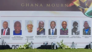 Row of head-and-shoulders portraits on a Ghana Mourns memorial banner at an outdoor ceremony with flowers in front.