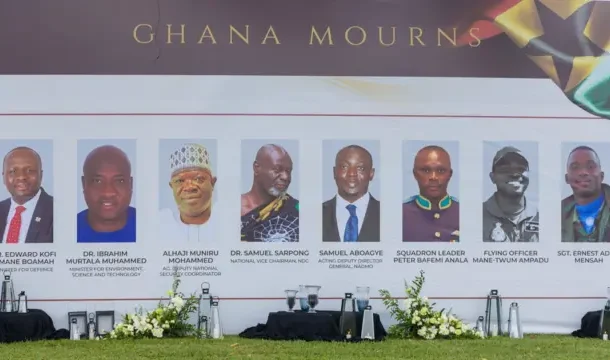 Row of head-and-shoulders portraits on a Ghana Mourns memorial banner at an outdoor ceremony with flowers in front.