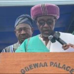 Man wearing traditional green attire and a purple hat speaks into a microphone at a wooden podium labeled 'GBEWAA PALACE', flanked by assistants.