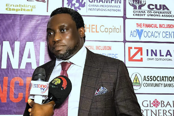 Man in a dark suit and red tie being interviewed, holding still while microphones are held up to him, with a sponsor-filled backdrop behind him.