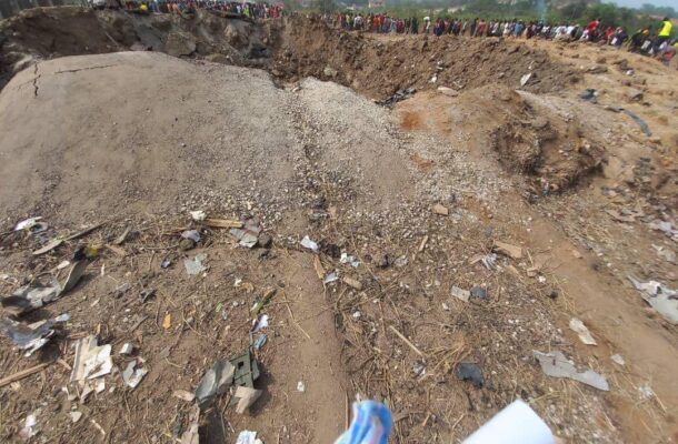 Crowd of people standing along the edge of a collapsed dirt slope with rubble and debris scattered across the ground behind a dirt path. A blue cloth is in the foreground.