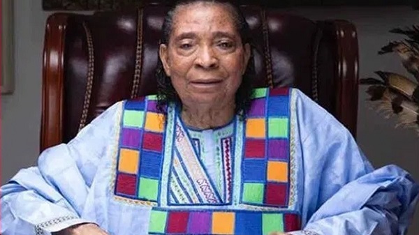 Older woman seated in a dark leather chair, wearing a light blue traditional dress with a colorful patchwork bib and sleeves.