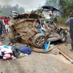 Wrecked car crushed with debris on a road, smoke in the air and a damaged bus in the background, with onlookers nearby.