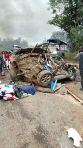 Wrecked car crushed with debris on a road, smoke in the air and a damaged bus in the background, with onlookers nearby.