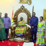 Group of officials and guests posing around a seated leader on an ornate throne in a ceremonial room, flags in view.