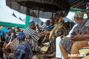 Senior Ghanaian dignitaries in ornate traditional robes shake hands beneath a large patterned umbrella at a public ceremony, surrounded by supporters and photographers.