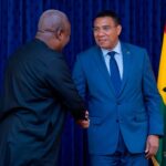 Two men in formal attire shake hands in front of a blue curtain, with a Ghana flag nearby, signaling a formal meeting or agreement.