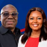 Two smiling adults standing side by side against a blue backdrop; man wears glasses and a black shirt, woman wears a white blazer over red top.