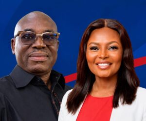 Two smiling adults standing side by side against a blue backdrop; man wears glasses and a black shirt, woman wears a white blazer over red top.