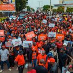 Large crowd of protesters in orange clothing fills a city street, holding signs and banners with police nearby on the left.