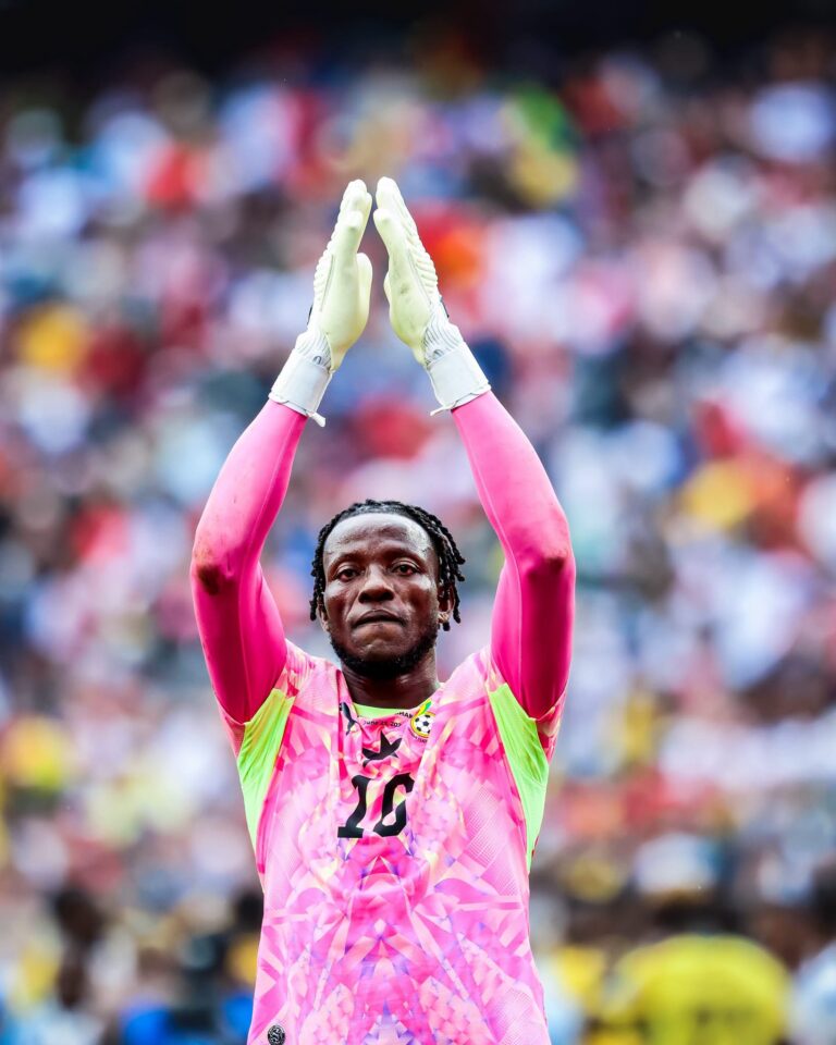 Soccer goalkeeper in a pink kit raises gloved hands together in celebration with a blurred colorful crowd behind.
