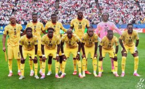 A soccer team in yellow uniforms poses for a pre-match group photo on a green field with a crowded stadium in the background.