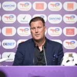 Man in a navy suit at a press conference with two microphones, seated behind a purple backdrop covered in sponsor logos and a soccer ball on the table nearby.