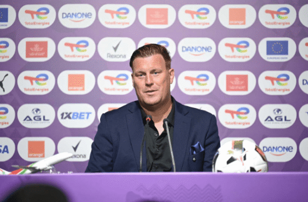 Man in a navy suit at a press conference with two microphones, seated behind a purple backdrop covered in sponsor logos and a soccer ball on the table nearby.
