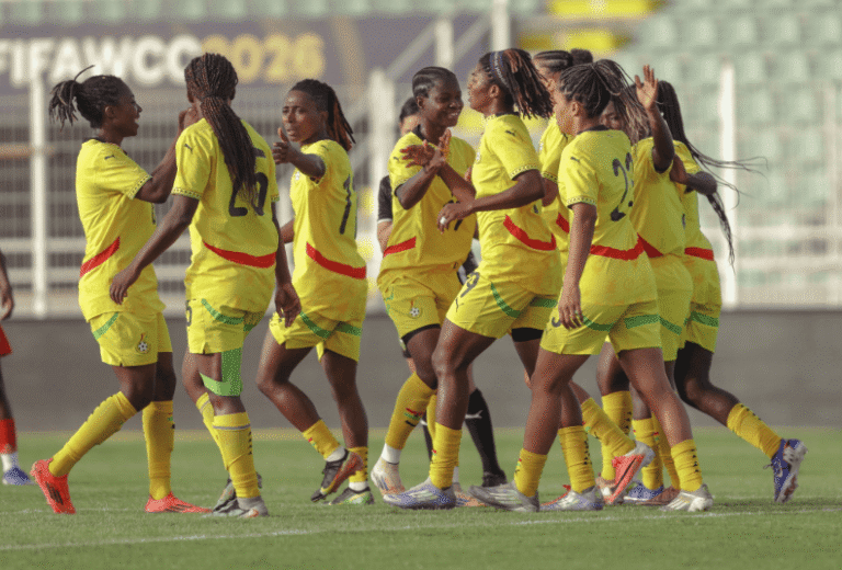 Young women in yellow soccer uniforms celebrate together on a pitch, arms raised and smiling after a match moment.