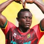 Soccer player in a red jersey making a heart shape with arms raised above his head on a sunny field.