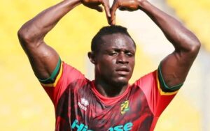 Soccer player in a red jersey making a heart shape with arms raised above his head on a sunny field.