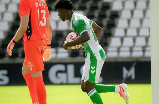 Soccer players in green/white and orange kits run near the goal; one player carries a ball as the orange-clad goalkeeper watches nearby.