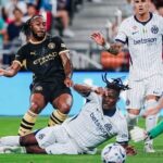 Soccer players collide for the ball on a green field: two Inter Milan players in white kits, one sliding, while a Manchester City player in black presses nearby.