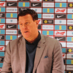 Man in a light gray blazer and navy sweater sits at a table during a press conference with sponsor logos behind him, speaking to the audience.