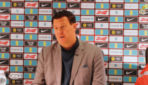 Man in a light gray blazer and navy sweater sits at a table during a press conference with sponsor logos behind him, speaking to the audience.