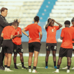 Coach or mentor addressing a group of female soccer players in red jerseys on a green field during a timeout.