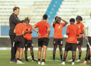 Coach or mentor addressing a group of female soccer players in red jerseys on a green field during a timeout.