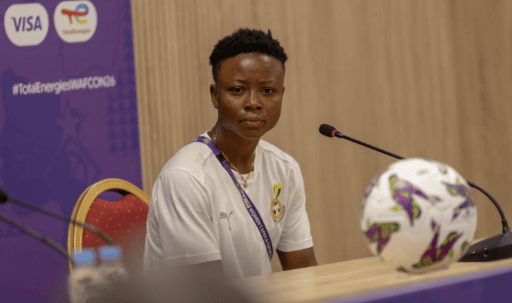 Female panelist in a white shirt sits at a conference table with a microphone, purple sponsor banner in the background.