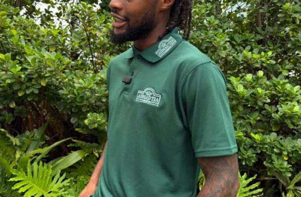 Side-profile of a man with braided hair and beard wearing a green Ahmad Tea polo in a lush garden setting.