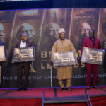 Five men stand on a stage, each holding a framed award plaque during a ceremony with a large backdrop featuring portraits and the words 'Presented to a Legend'.