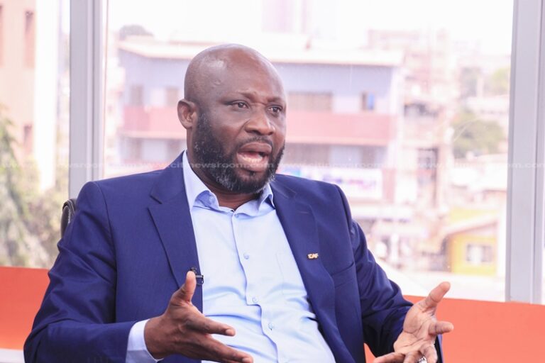 African man in a blue suit and light blue shirt speaks and gestures with his hands during a discussion in a bright office setting.