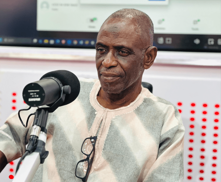 Man in a light patterned shirt sits at a microphone in a radio studio, wearing glasses resting on his chest, with a computer screen and red soundproof panels in the background.