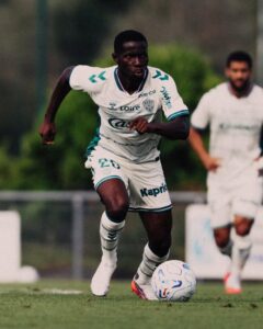 Soccer player in a white kit with teal trim dribbling a ball on a grassy field during a match.