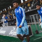 Soccer player in a blue training kit with number 5 walking along the sideline near the dugout.
