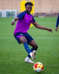 Soccer player in a purple training jersey and blue shorts kicks a ball on a green field during practice, focusing on footwork.