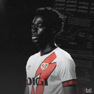 Portrait of a young Black soccer player in a white jersey with bold red graphic, captain's armband, against a dark chalkboard background.