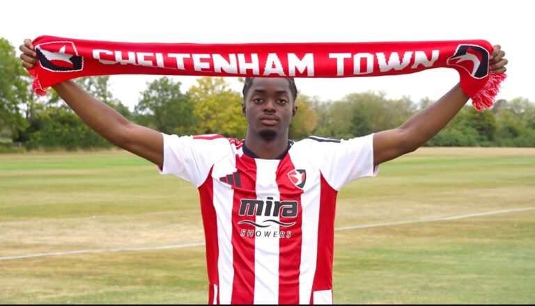 Soccer player in a red-and-white striped jersey raises a red Cheltenham Town scarf above his head on a grassy field.