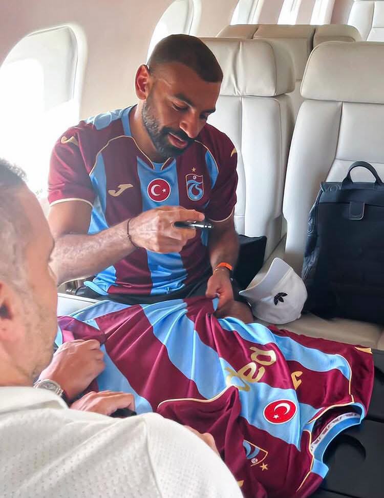 Footballer in a maroon-and-blue jersey sits in a plane seat, signing memorabilia with a pen while another person watches from the foreground.