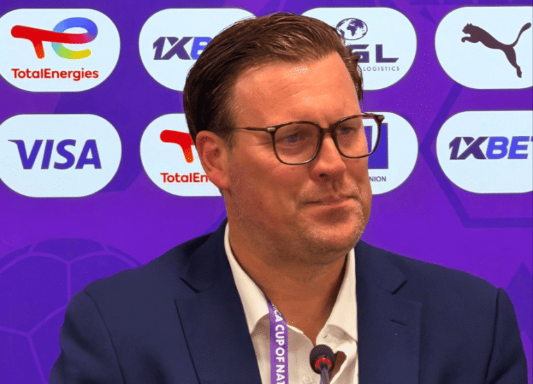 Man in a navy suit and glasses speaking into a microphone at a conference, with sponsor logos on a purple backdrop.