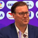 Man in a dark suit with glasses speaks at a conference microphone, backdrop filled with sponsor logos.