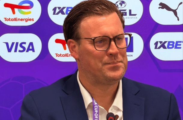 Man in a dark suit with glasses speaks at a conference microphone, backdrop filled with sponsor logos.