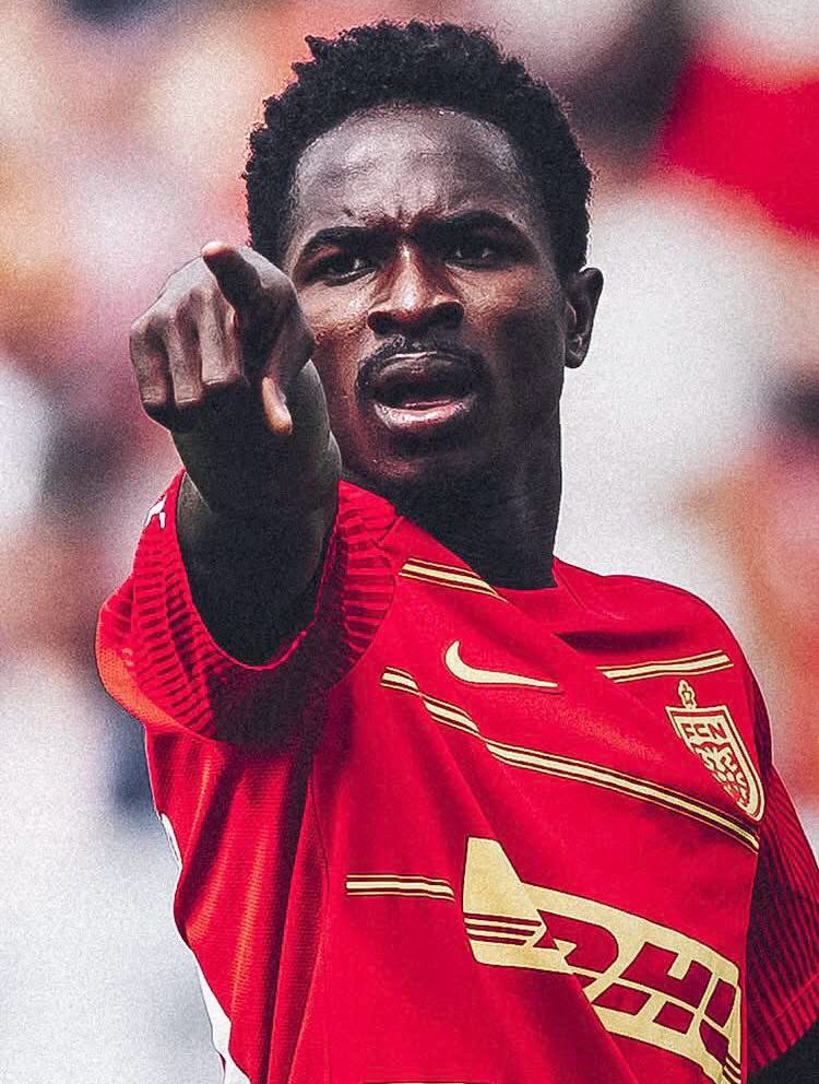 Soccer player in a red jersey points toward the camera during a match, stadium blurred in the background.