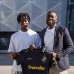 Two men stand outside a modern building, holding up a black soccer jersey with yellow trim and the sponsor 'truecaller'.