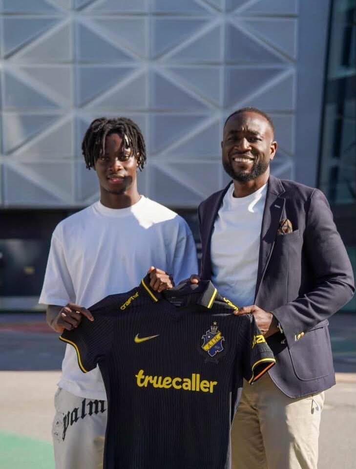 Two men stand outside a modern building, holding up a black soccer jersey with yellow trim and the sponsor 'truecaller'.