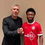 Two men shake hands and smile for the camera; one in a black blazer, the other in a red sports jersey, against a beige wall background.