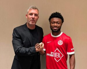 Two men shake hands and smile for the camera; one in a black blazer, the other in a red sports jersey, against a beige wall background.