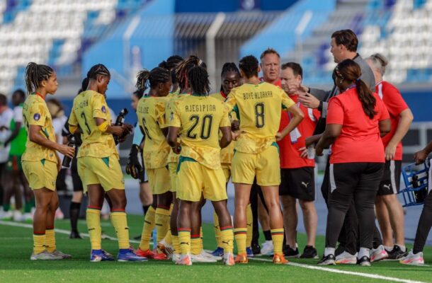 Female soccer players in yellow uniforms form a huddle on the green field as coaches in red shirts talk to them.