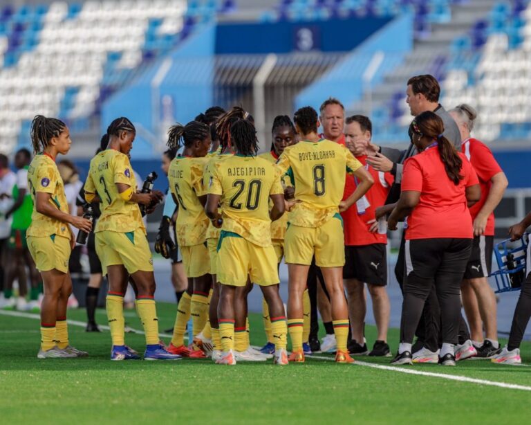 Female soccer players in yellow uniforms form a huddle on the green field as coaches in red shirts talk to them.