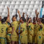 Women’s soccer team in yellow uniforms huddle and celebrate on the field, arms raised, players cheering together in a stadium.