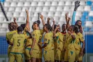 Women’s soccer team in yellow uniforms huddle and celebrate on the field, arms raised, players cheering together in a stadium.